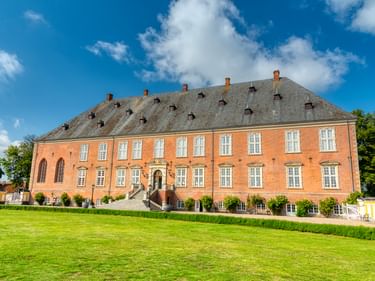 Valdemars Slot, ein großes Barockschloss aus rotem Backstein mit grauem Mansardendach auf der Insel Tåsinge. Das symmetrische Gebäude hat weiße Fenster und eine zentrale Treppe, umgeben von gepflegten Rasenflächen.