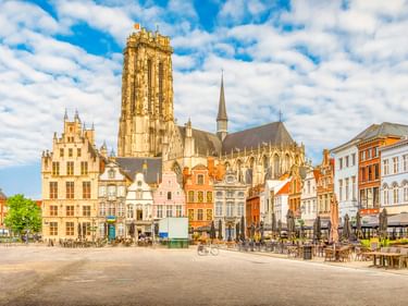Marktplatz in Mechelen mit dem Turm der St.-Rombouts-Kathedrale über bunten historischen Gebäuden. Blauer Himmel mit weißen Wolken.
