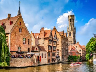 Historische Kanalszene in Brügge mit mittelalterlichen Backsteinhäusern und dem hohen Belfried. Touristenboot auf dem Wasser, Menschen am Kai.