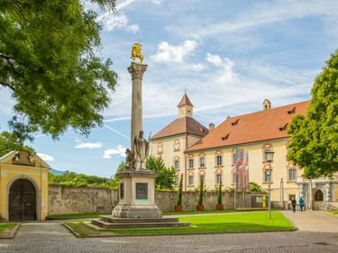 Hofburg in Brixen mit gelber Fassade, orangefarbenem Ziegeldach und Innenhof mit hoher Säule und goldener Statue, eingerahmt von grünen Bäumen.