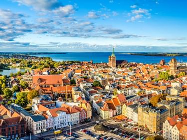 Luftaufnahme der Altstadt Stralsund mit roten Ziegeldächern, Kirchtürmen und der blauen Ostsee im Hintergrund unter bewölktem Himmel.