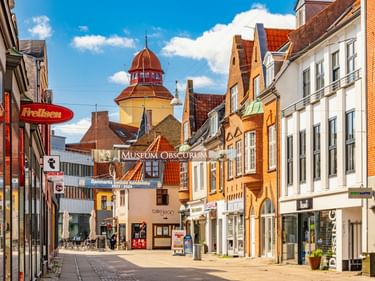 Bunte Fußgängerzone in Nykøbing Falster mit historischen Gebäuden, Geschäften und gelbem Turm mit roter Kuppel unter blauem Himmel.