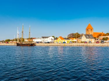 Hafenblick auf Stege mit bunten Gebäuden am Wasser, einem historischen Segelschiff und einer Kirche mit orangem Turm unter blauem Himmel.