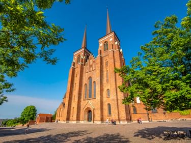 Roskilder Dom aus rotem Backstein mit zwei Türmen, eingerahmt von grünen Bäumen unter blauem Himmel. Besucher sitzen auf dem Kopfsteinpflaster.