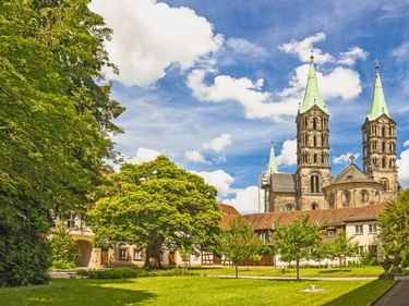 Bamberg Cathedral with four towers topped by green copper spires, surrounded by historic buildings and a green lawn with trees under a blue sky.
