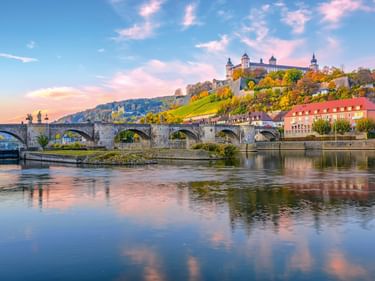 Marienberg Fortress on a hilltop overlooking the Old Main Bridge in Würzburg at sunset. The stone bridge spans the Main River with reflections in calm water.