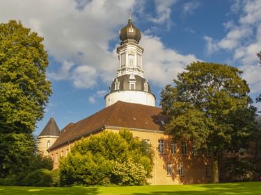 Schloss Jever mit gelber Fassade und verziertem weißen Turm mit dunkler Kuppel, umgeben von grüner Wiese und alten Bäumen unter blauem Himmel.