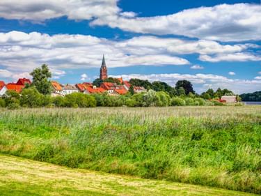 Blick auf Röbel an der Müritz mit rotgedeckten Häusern und Kirchturm, grüne Wiese und Schilf im Vordergrund, See und blauer Himmel mit Wolken.