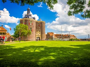 Radfahrer auf grüner Wiese vor historischem Rundturm in Enkhuizen. Traditionelle niederländische Häuser mit orangen Dächern unter blauem Himmel.
