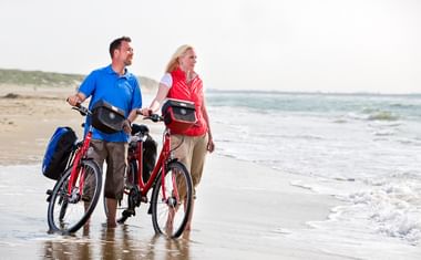 Mann und Frau mit roten Tourenrädern am Wassersaum eines Strandes in Zeeland, mit Dünen und Meer im Hintergrund.