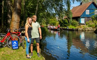 Paar mit Fahrrädern am Kanal im Spreewald. Traditionelles blaues Haus mit rotem Dach und Touristenboot im Hintergrund sichtbar.