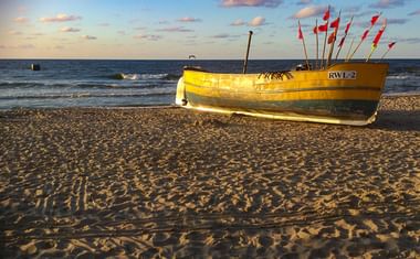 Gelbes Fischerboot mit Kennzeichen RWL-2 und roten Flaggen am Sandstrand bei Sonnenuntergang. Ostseewellen und blauer Himmel mit rosa Wolken im Hintergrund.