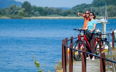 Zwei Radfahrer mit roten Fahrrädern auf einem Uferweg am Überlinger See, Bodensee. Blaues Wasser, Segelboote und grüne Hügel im Hintergrund.