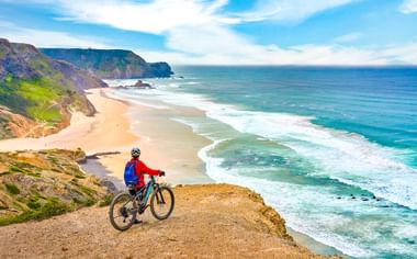 Radfahrer in roter Jacke auf Klippe mit Blick auf Sandstrand und türkisfarbenen Atlantik bei Sagres. Dramatische Küstenklippen rahmen die Bucht.