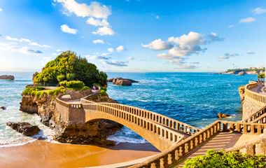 Steinerne Fußgängerbrücke mit Balustraden zu einer grünen Felseninsel in Biarritz. Blaues Meer, Sandstrand und Küstengebäude unter sonnigem Himmel.