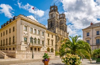 Kathedrale Sainte-Croix in Orléans mit gotischen Zwillingstürmen hinter einem cremefarbenen Gebäude. Palmen und Blumen schmücken den sonnigen Platz.
