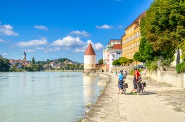 Cyclists on riverside path along the Inn River in Passau with historic tower, colorful buildings, and church spire under blue sky.