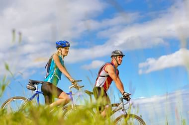 Two cyclists with helmets and backpacks riding mountain bikes through tall grass under a blue sky with white clouds.