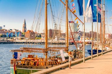 Traditionelle hölzerne Segelschiffe mit hohen Masten am Kieler Hafen. Kirchturm und Stadtgebäude im Hintergrund unter blauem Himmel.