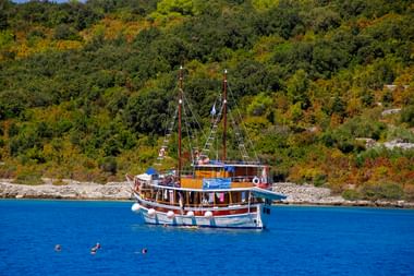 MS Orkan, ein traditionelles Holzschiff mit zwei Masten, ankert in blauem Wasser nahe einer bewaldeten Küste. Schwimmer im Wasser sichtbar.