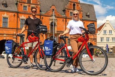 Zwei Radfahrer mit roten Mieträdern mit blauen Taschen stehen vor einem historischen roten Backsteingebäude am Marktplatz in Brunsbüttel.