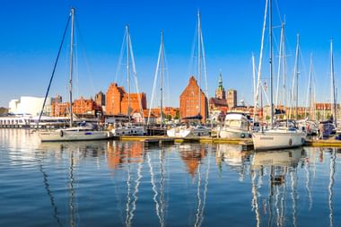 Marina in Stralsund mit Segelbooten im ruhigen Wasser. Historische Backsteingebäude und Kirchtürme säumen die Uferpromenade unter blauem Himmel.