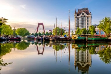 Historische Schiffe im Alten Hafen Rotterdam mit Willemsbrücke und Witte Huis, die sich im ruhigen Wasser bei Sonnenuntergang spiegeln.