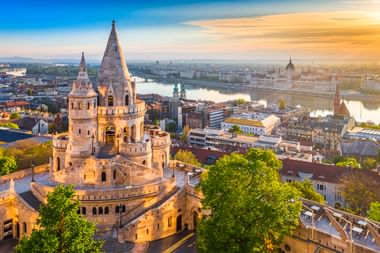 Fisherman's Bastion towers in Budapest with the Danube River and Parliament Building visible in the background at sunset.