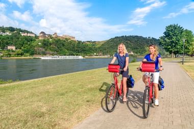Zwei Radfahrerinnen mit roten Taschen auf Rheinuferweg. Flusskreuzfahrtschiff und Burg auf Hügel im Hintergrund unter blauem Himmel.