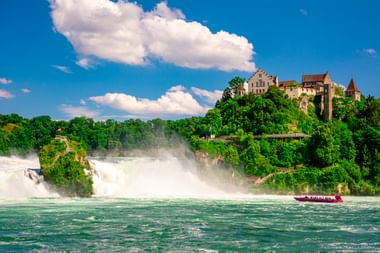 Rheinfall mit kraftvoll herabstürzendem Wasser und Gischt. Ein rotes Touristenboot im türkisfarbenen Fluss. Schloss Laufen auf grünem Hügel.