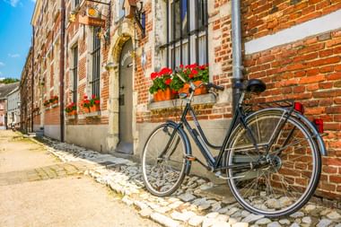 Blaues Fahrrad auf Kopfsteinpflasterstraße vor Backsteingebäude mit roten Geranien in Blumenkästen in Herentals, Belgien.