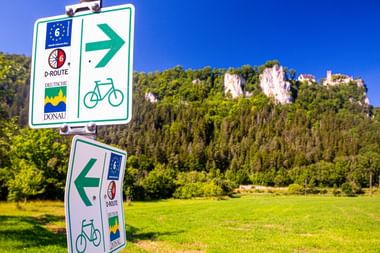 Donauradweg-Wegweiser mit Fahrradsymbolen nach links und rechts, mit Schloss Werenwag auf bewaldeten Klippen im Hintergrund.