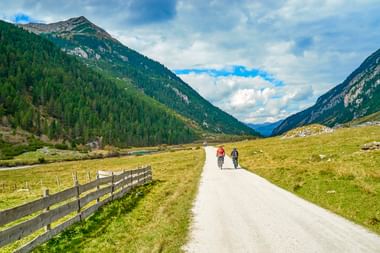 Radfahrer und Fußgänger auf asphaltiertem Weg durch alpines Tal bei Krimml. Holzzaun säumt die Route mit grünen Wiesen und bewaldeten Bergen.