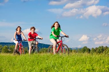 Three children cycling on red bicycles through a lush green meadow under a blue sky with white clouds. Forested hills visible in background.