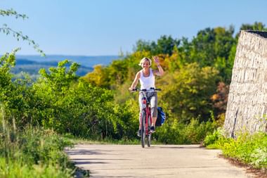 Radfahrerin winkt beim Fahren auf asphaltiertem Weg durch Zellingen Weinberge. Grüne Reben und Bäume säumen die Route unter blauem Himmel.