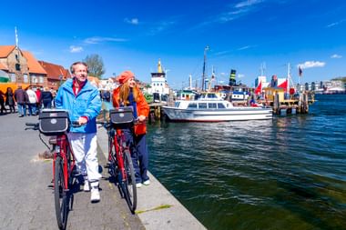 Zwei Radfahrer mit roten Rädern am Flensburger Hafen. Dahinter liegen Boote, bunte Flaggen und historische Gebäude unter blauem Himmel.
