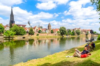 Zwei Radfahrer ruhen auf der Wiese an der Donau in Ulm. Historische Skyline mit Ulmer Münster und bunten Gebäuden spiegelt sich im Wasser.