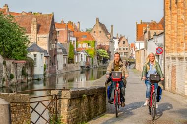 Zwei Radfahrer mit roten Rädern überqueren eine Steinbrücke über einen Kanal in Brügge, mit mittelalterlichen Backsteingebäuden und orangefarbenen Ziegeldächern im Hintergrund.