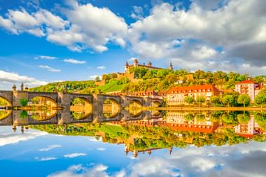 Steinbrücke über den Main spiegelt die Festung Marienberg auf dem Hügel in Würzburg. Gebäude mit roten Dächern säumen das Flussufer unter blauem Himmel.
