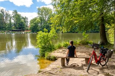Person sitzt auf Holzbank am sandigen Ufer des Rosensees in Schwentinental, rotes Fahrrad steht unter großem Baum.