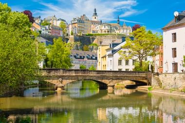Steinbrücke über ruhigem Fluss in Luxemburg-Stadt mit bunten historischen Gebäuden am Hang, grünen Bäumen und blauem Himmel.