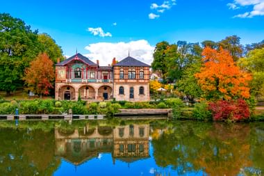 Verziertes historisches Gebäude mit Arkadengang spiegelt sich im Teich, umgeben von bunten Herbstbäumen in Sarreguemines unter blauem Himmel.