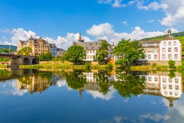 Bernkastel-Kues am Moselufer mit historischen Gebäuden und Kirchtürmen, die sich im ruhigen Fluss spiegeln, unter blauem Himmel mit weißen Wolken.