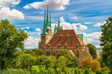 Erfurter Dom mit markanten grünen Kupfertürmen und rotem Ziegeldach, umgeben von grünen Bäumen unter blauem Himmel mit weißen Wolken.