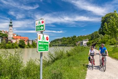 Zwei Radfahrer auf einem asphaltierten Weg entlang eines grünen Flusses. Radwegschilder weisen zum Tauernradweg und Salzburg. Historische Stadt mit Kirchturm sichtbar.