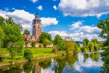 St. Crucis Kirche mit hohem Steinturm neben einem Fluss in Bad Sooden-Allendorf, Hessen. Grüne Wiesen und Bäume umgeben die Kirche unter blauem Himmel.
