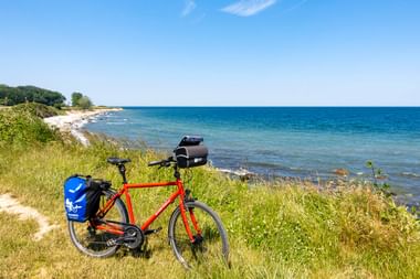 Rotes Reiserad mit blauen Packtaschen auf grasigem Küstenweg mit Blick auf die Ostsee auf Fehmarn unter blauem Himmel.