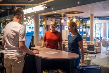 Lächelndes Paar mit Gepäck an der Hotelrezeption im Gespräch mit Mitarbeiter. Moderne Lobby mit Loungebereich und großen Fenstern im Hintergrund.