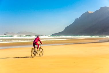 Radfahrer in pinker Jacke fährt auf goldenem Sand am Strand Playa de Famara mit dramatischen Klippen und türkisfarbenen Meereswellen im Hintergrund.