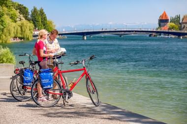 Zwei Radfahrer mit Rädern lesen eine Karte am Seerheinufer in Konstanz. Historischer Turm und Brücke über dem türkisfarbenen Wasser.